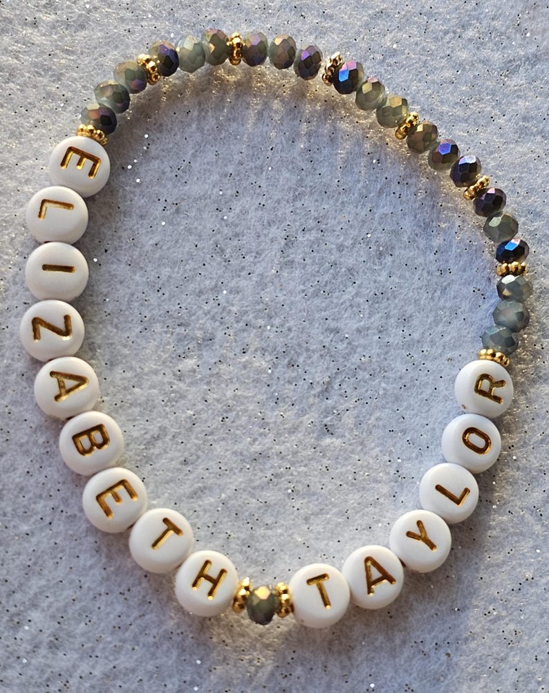May include: A beaded bracelet featuring white circular beads with gold lettering spelling out ELIZABETH TAYLOR, interspersed with small gold-toned spacers and a section of faceted, dark grey, translucent beads arranged in a circular pattern.