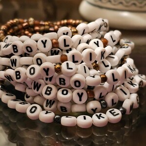 May include: A stack of beaded bracelets featuring white round beads with black letters spelling out COWBOY LIKE ME, interspersed with small metallic bronze and dark grey spherical beads. The bracelets are arranged in a circular, layered formation.