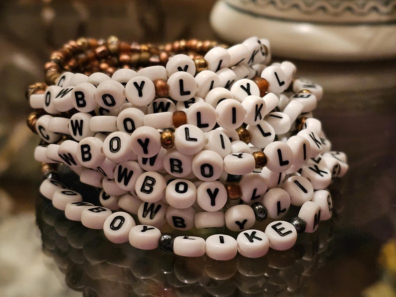 May include: A stack of beaded bracelets featuring white round beads with black letters spelling out COWBOY LIKE ME, interspersed with small metallic bronze and dark grey spherical beads. The bracelets are arranged in a circular, layered formation.