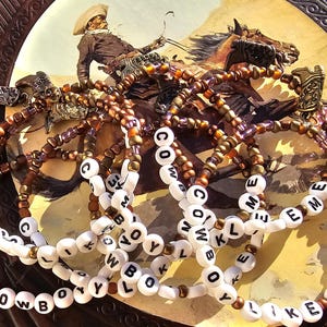 May include: A collection of beaded bracelets featuring metallic bronze and purple seed beads, white letter beads spelling out the phrase COWBOY LIKE ME, and small antique-style metal cowboy boot charms attached to each individual bracelet strand.