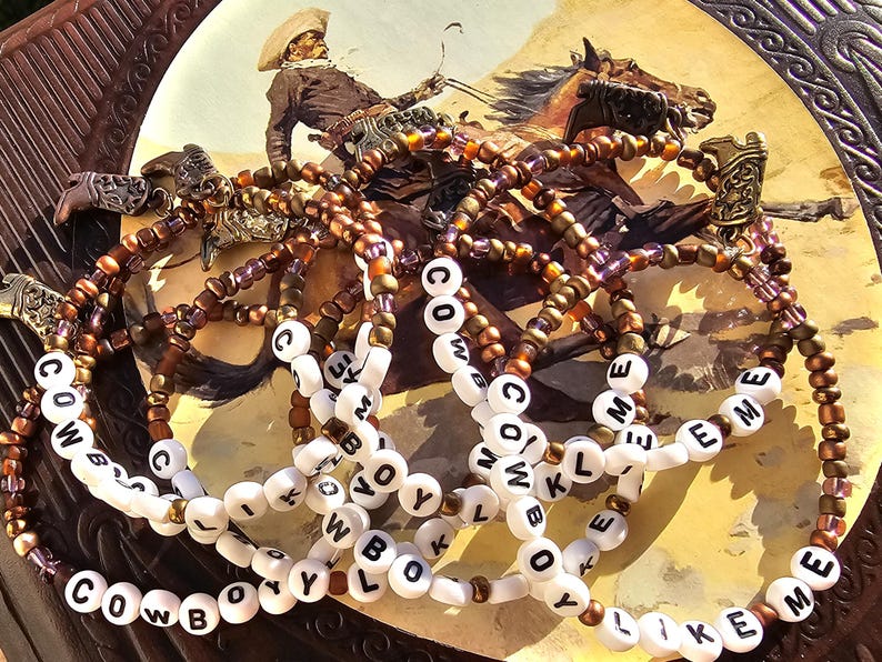 May include: A collection of beaded bracelets featuring metallic bronze and purple seed beads, white letter beads spelling out the phrase COWBOY LIKE ME, and small antique-style metal cowboy boot charms attached to each individual bracelet strand.