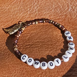 May include: A beaded bracelet featuring a mix of metallic bronze and brown beads, white letter beads spelling out COWBOY LIKE ME, and a small gold-toned metal cowboy boot charm attached to the side of the circular beaded strand.