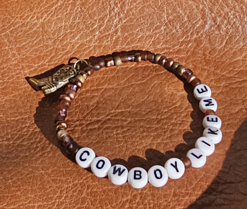 May include: A beaded bracelet featuring a mix of metallic bronze and brown beads, white letter beads spelling out COWBOY LIKE ME, and a small gold-toned metal cowboy boot charm attached to the side of the circular beaded strand.