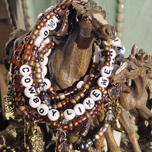 May include: A stack of beaded bracelets featuring white letter beads spelling COWBOY LIKE ME, interspersed with metallic bronze, copper, and black beads. The bracelets include small dangling charms shaped like cowboy boots in various metallic finishes.