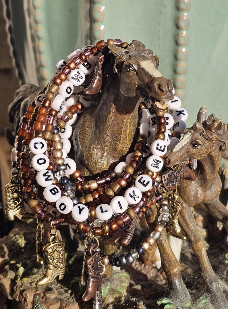 May include: A stack of beaded bracelets featuring white letter beads spelling COWBOY LIKE ME, interspersed with metallic bronze, copper, and black beads. The bracelets include small dangling charms shaped like cowboy boots in various metallic finishes.