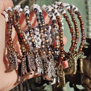 May include: A collection of beaded bracelets featuring white letter beads spelling COWBOY, interspersed with metallic bronze, copper, and silver-toned seed beads. Each bracelet includes a small metal charm shaped like a western cowboy boot.