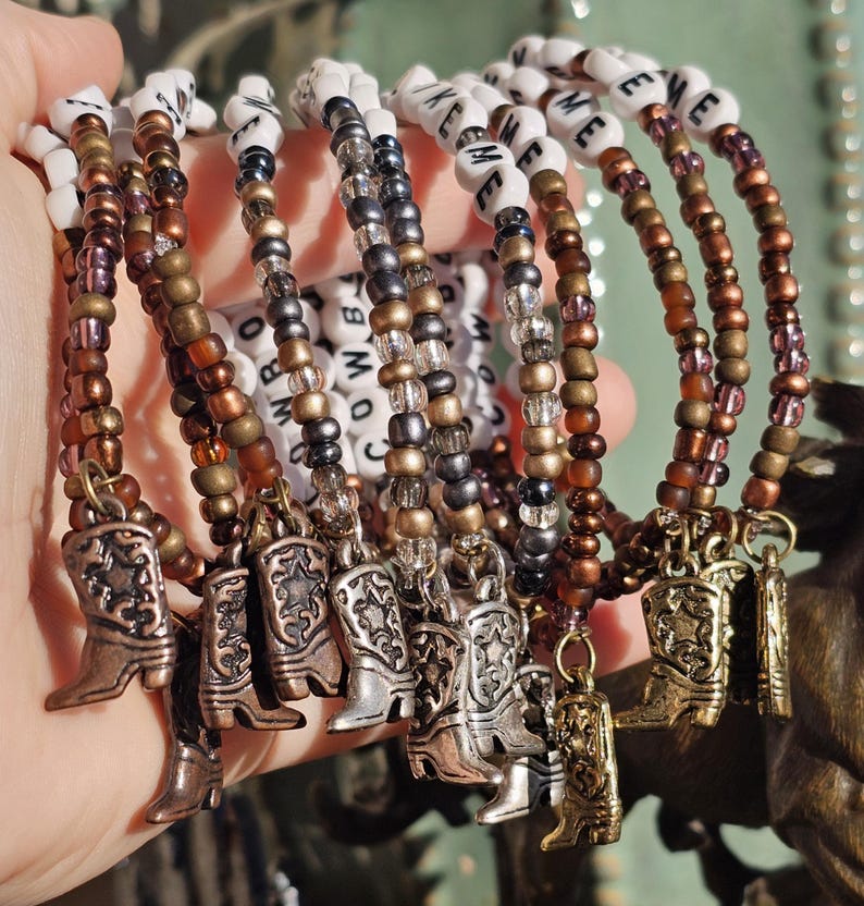 May include: A collection of beaded bracelets featuring white letter beads spelling COWBOY, interspersed with metallic bronze, copper, and silver-toned seed beads. Each bracelet includes a small metal charm shaped like a western cowboy boot.