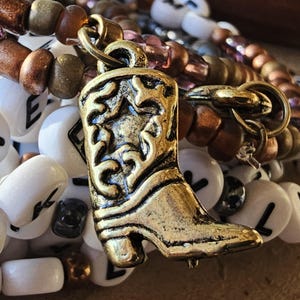 May include: A bracelet featuring a gold-toned metal cowboy boot charm with star detailing, layered with white alphabet beads displaying various black letters and a strand of metallic bronze and translucent purple glass seed beads.