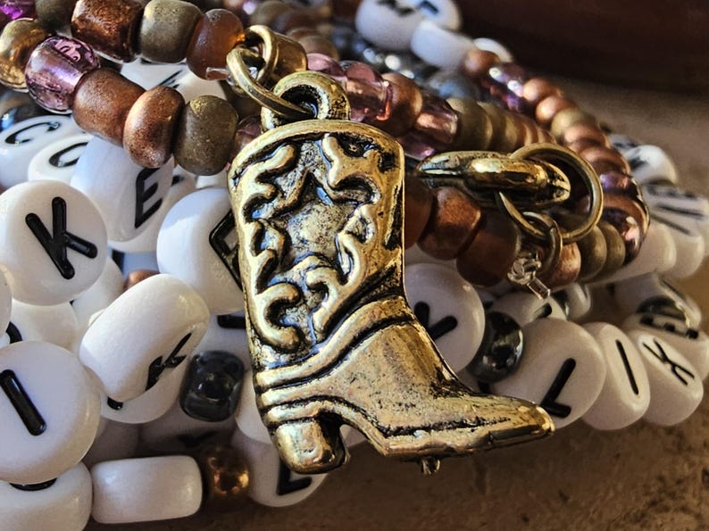 May include: A bracelet featuring a gold-toned metal cowboy boot charm with star detailing, layered with white alphabet beads displaying various black letters and a strand of metallic bronze and translucent purple glass seed beads.