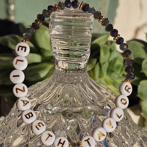 May include: A beaded bracelet featuring white circular beads with gold lettering that spells out ELIZABETH TAYLOR. The bracelet is accented with dark, faceted, iridescent beads and small gold-toned spacers, creating a circular design.