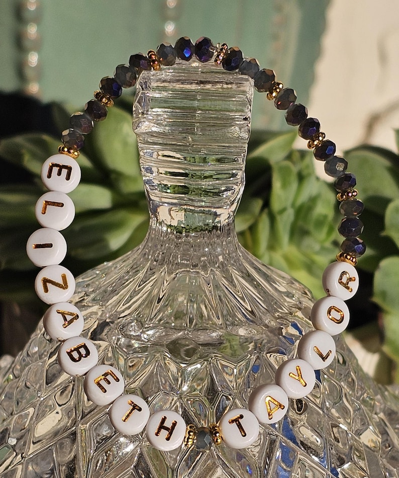May include: A beaded bracelet featuring white circular beads with gold lettering that spells out ELIZABETH TAYLOR. The bracelet is accented with dark, faceted, iridescent beads and small gold-toned spacers, creating a circular design.