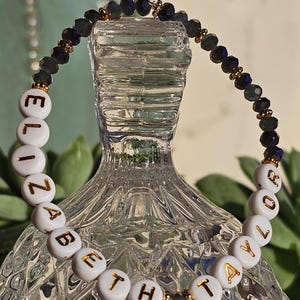 May include: A beaded bracelet featuring white circular beads with gold lettering that spells out ELIZABETH TAYLOR. The bracelet is accented with dark, faceted, iridescent beads and small gold-toned spacers, creating a contrast between the letters and beads.