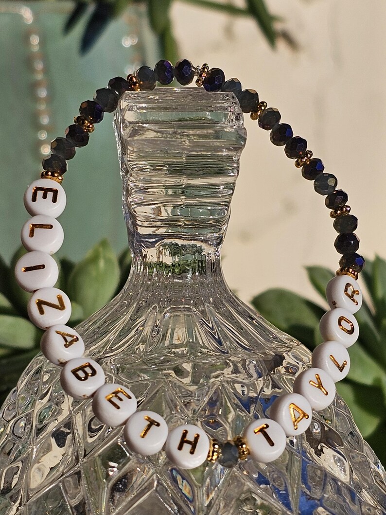 May include: A beaded bracelet featuring white circular beads with gold lettering that spells out ELIZABETH TAYLOR. The bracelet is accented with dark, faceted, iridescent beads and small gold-toned spacers, creating a contrast between the letters and beads.