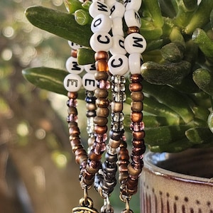May include: Three beaded bracelets featuring white letter beads spelling COW LIKE ME, interspersed with metallic bronze, silver, and translucent purple seed beads. Each bracelet includes a dangling metal cowboy boot charm with star detail.
