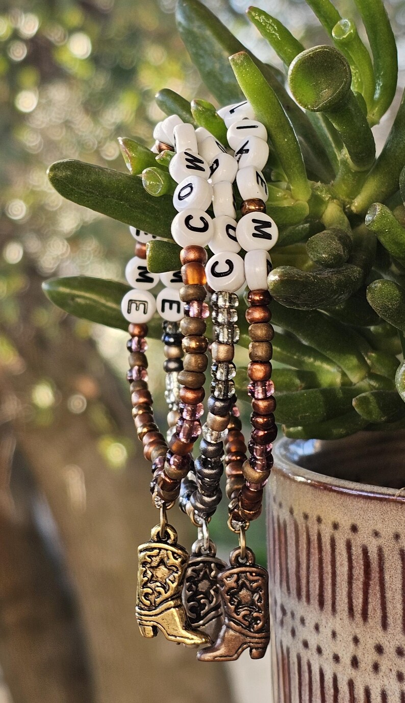 May include: Three beaded bracelets featuring white letter beads spelling COW LIKE ME, interspersed with metallic bronze, silver, and translucent purple seed beads. Each bracelet includes a dangling metal cowboy boot charm with star detail.