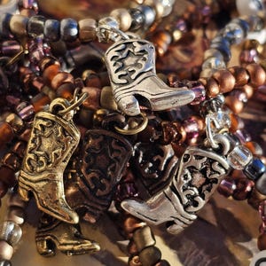 May include: A collection of beaded bracelets featuring metallic cowboy boot charms. The bracelets are composed of small, round beads in shades of bronze, copper, silver, and translucent purple. The charms are detailed with star patterns.