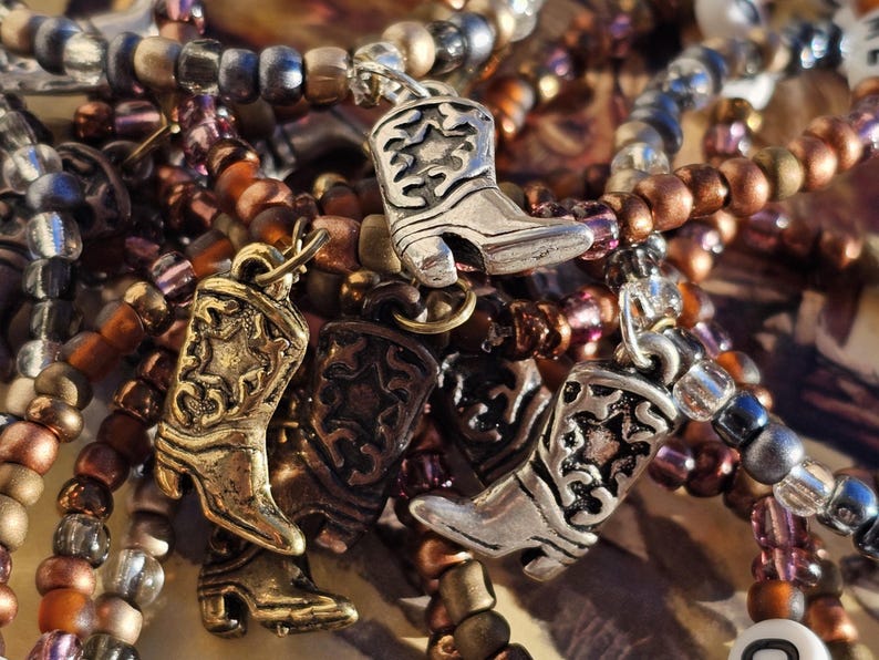 May include: A collection of beaded bracelets featuring metallic cowboy boot charms. The bracelets are composed of small, round beads in shades of bronze, copper, silver, and translucent purple. The charms are detailed with star patterns.