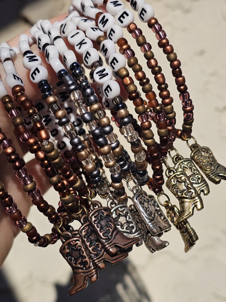 May include: A collection of beaded bracelets featuring white letter beads spelling LIKE ME, interspersed with metallic bronze, copper, and silver-toned seed beads. Each bracelet is finished with a small metal cowboy boot charm pendant.