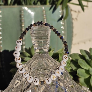 May include: A beaded bracelet featuring white circular beads with gold lettering spelling out ELIZABETH TAYLOR, interspersed with small dark faceted beads and tiny gold-toned metallic spacers arranged in a circular pattern.