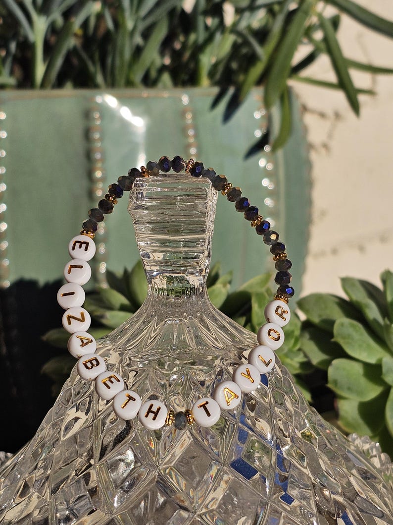 May include: A beaded bracelet featuring white circular beads with gold lettering spelling out ELIZABETH TAYLOR, interspersed with small dark faceted beads and tiny gold-toned metallic spacers arranged in a circular pattern.