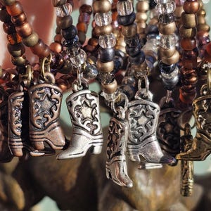 May include: A collection of beaded bracelets featuring metallic cowboy boot charms. The bracelets are constructed with small, round beads in shades of brown, bronze, and clear glass. Each bracelet includes a dangling metal boot charm with a star.