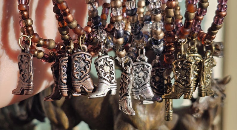 May include: A collection of beaded bracelets featuring metallic cowboy boot charms. The bracelets are constructed with small, round beads in shades of brown, bronze, and clear glass. Each bracelet includes a dangling metal boot charm with a star.