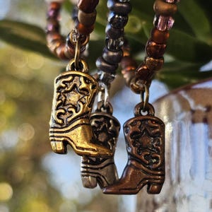 May include: Three beaded bracelets featuring metallic cowboy boot charms. The charms are finished in gold, silver, and copper tones, each displaying an embossed star and scrollwork pattern. The beads are arranged in brown, grey, and bronze hues.