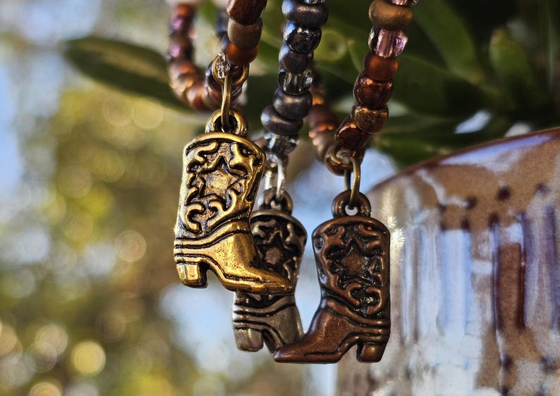 May include: Three beaded bracelets featuring metallic cowboy boot charms. The charms are finished in gold, silver, and copper tones, each displaying an embossed star and scrollwork pattern. The beads are arranged in brown, grey, and bronze hues.