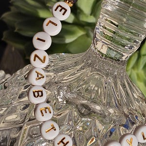 May include: A beaded bracelet featuring white circular beads with gold lettering spelling out ELIZABETH TAYLOR. The design includes dark grey faceted beads and small gold-toned spacers, creating a contrast between the white lettered beads and the dark accents.