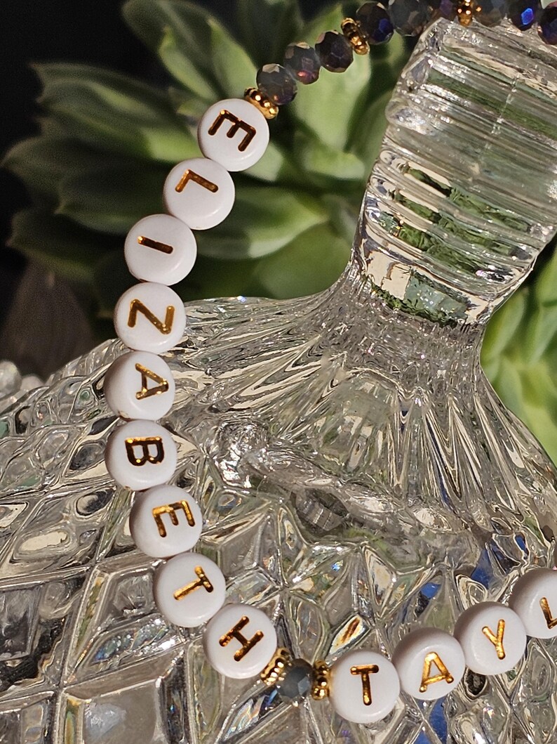 May include: A beaded bracelet featuring white circular beads with gold lettering spelling out ELIZABETH TAYLOR. The design includes dark grey faceted beads and small gold-toned spacers, creating a contrast between the white lettered beads and the dark accents.