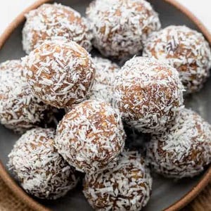 Op de afbeelding: Een bord met energieballen, bedekt met geraspte kokos. De ballen zijn bruin en rond, en de kokos is wit. Het grijze bord heeft een bruine rand en ligt op een jute doek.