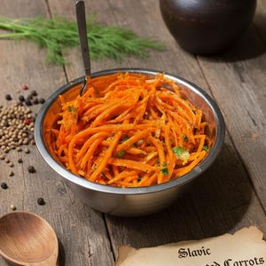May include: A stainless steel bowl filled with orange shredded carrots, a wooden spoon, and a small dark brown ceramic pot sit on a rustic wooden surface. The image includes a sprig of dill, peppercorns, and a parchment label that reads "Slavic Fermented Carrots - Probiotic Delight - Hand-Crafted."