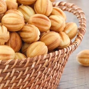 Op de afbeelding: Een rieten mand vol goudbruine, walnootvormige koekjes. De koekjes hebben een textuur en zijn gerangschikt in een rustieke mand, met een paar koekjes verspreid. De afbeelding roept een gevoel van warmte en zelfgemaakte lekkernijen op.