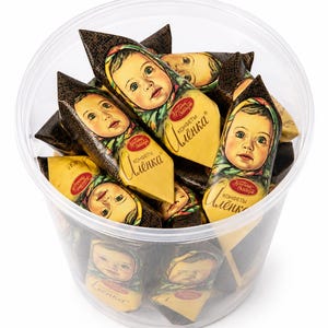 May include: A clear plastic container filled with individually wrapped candies. Each candy wrapper features a portrait of a baby with a headscarf. The wrappers are primarily yellow and brown, with text in a foreign language. The candies are a traditional Russian confection.