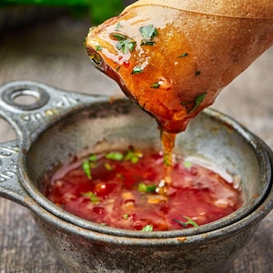 Puede incluir: Primer plano de un rollito de primavera sumergiéndose en un cuenco metálico pequeño con salsa roja. La salsa está salpicada de hierbas verdes. El rollito de primavera es dorado.