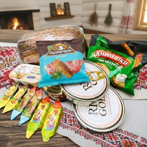 Könnte beinhalten: Eine Sammlung verpackter Lebensmittel, darunter ein Brotlaib, eine Tüte Chips mit dem Text "BEKOH" und mehrere Dosen "RI GOLD". Weitere Artikel sind verpackte Süßigkeiten und ein Behälter mit Aufstrich. Die Artikel sind auf einem Tisch mit einem gemusterten Tuch angeordnet.