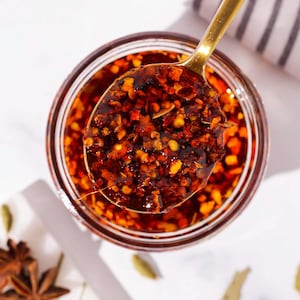 Puede incluir: Un tarro de aceite de chile con una cuchara dorada sacando una porción. El aceite es de color rojo intenso, lleno de copos y semillas de chile visibles. La cuchara está llena de aceite, mostrando su textura e ingredientes. La imagen está tomada desde arriba.