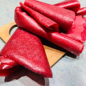 Puede incluir: Primer plano de rollos y láminas de cuero de fruta rojo brillante sobre una pequeña tabla de cortar de madera. El cuero de fruta tiene una superficie texturizada y está dispuesto de forma atractiva, sugiriendo un producto artesanal.