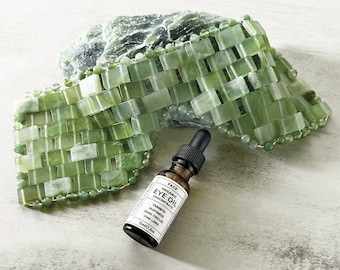 May include: Green jade gua sha tool with a small brown glass bottle labelled "FACE ORGANIC EYE OIL CONCENTRATE" with the text "TARGETS: PUFFINESS, DARK CIRCLES, FINE LINES 15ML/0.5oz"