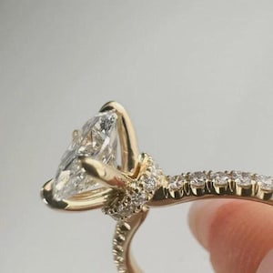 Puede incluir: Primer plano de un anillo de compromiso dorado con un diamante en forma de pera. La banda está adornada con diamantes más pequeños, creando un halo alrededor de la piedra principal. El anillo se muestra sobre un fondo neutro.
