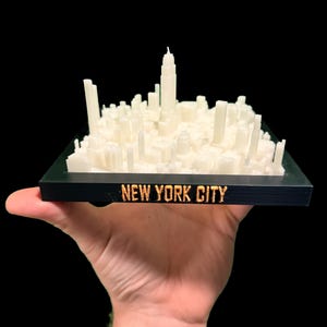 May include: A 3D-printed model of the New York City skyline. The model is off-white and sits on a black base with "NEW YORK CITY" in gold lettering. The model is held against a black background.