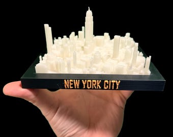 Puede incluir: Un modelo impreso en 3D del horizonte de la ciudad de Nueva York. El modelo es de color blanquecino y se asienta sobre una base negra con la inscripción "NEW YORK CITY" en letras doradas. El modelo se sostiene sobre un fondo negro.