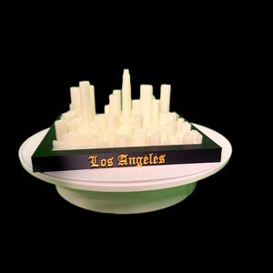 Puede incluir: Modelo impreso en 3D del horizonte de Los Ángeles. El modelo presenta edificios rectangulares blancos sobre una base negra, con "Los Ángeles" en letras doradas. El modelo se asienta sobre una base circular blanca.