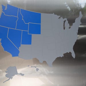 May include: A metallic surface featuring a map of the United States. The western region of the country is highlighted in a solid blue color, while the remaining states and the states of Alaska and Hawaii are depicted in a neutral gray color.
