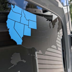 May include: A vinyl decal of the United States map applied to a window. The western portion of the contiguous United States is highlighted in bright blue, while the remainder of the country, including Alaska and Hawaii, is in matte black.