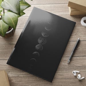 May include: A rectangular black notebook featuring a vertical sequence of white moon phases printed on the front cover. The cover has a smooth, reflective finish, and the notebook is positioned at an angle on a light-colored wooden surface.