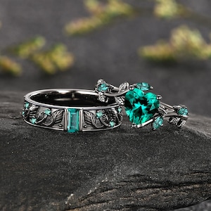 May include: A black metal ring set featuring a band with an emerald-cut green stone and a second band with a heart-shaped green stone. Both rings are decorated with intricate vine and leaf motifs, accented with small, round green stones.