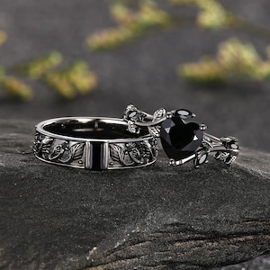 May include: A set of two black rings. One ring features a band with an embossed leaf pattern. The second ring has a heart-shaped black gemstone centerpiece held by a setting designed to resemble intertwined vines with small black accent stones.