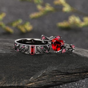 May include: A set of two black metal rings featuring intricate leaf and vine engravings. One ring has a central baguette-cut red gemstone, while the other features a prominent heart-shaped red gemstone, both accented with small round red stones.