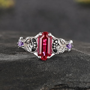 May include: A silver-toned ring featuring a central rectangular red gemstone. The band is decorated with intricate metalwork, small black circular stones, and two small purple round gemstones positioned on the outer sides of the setting.